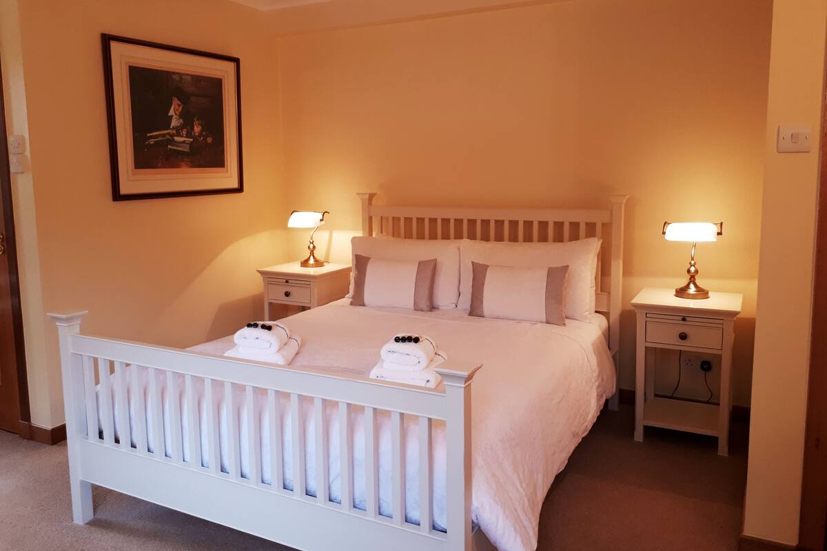 A neatly made bed with a white headboard and footboard, flanked by two bedside tables with lamps. A framed picture hangs on the wall above the bed.