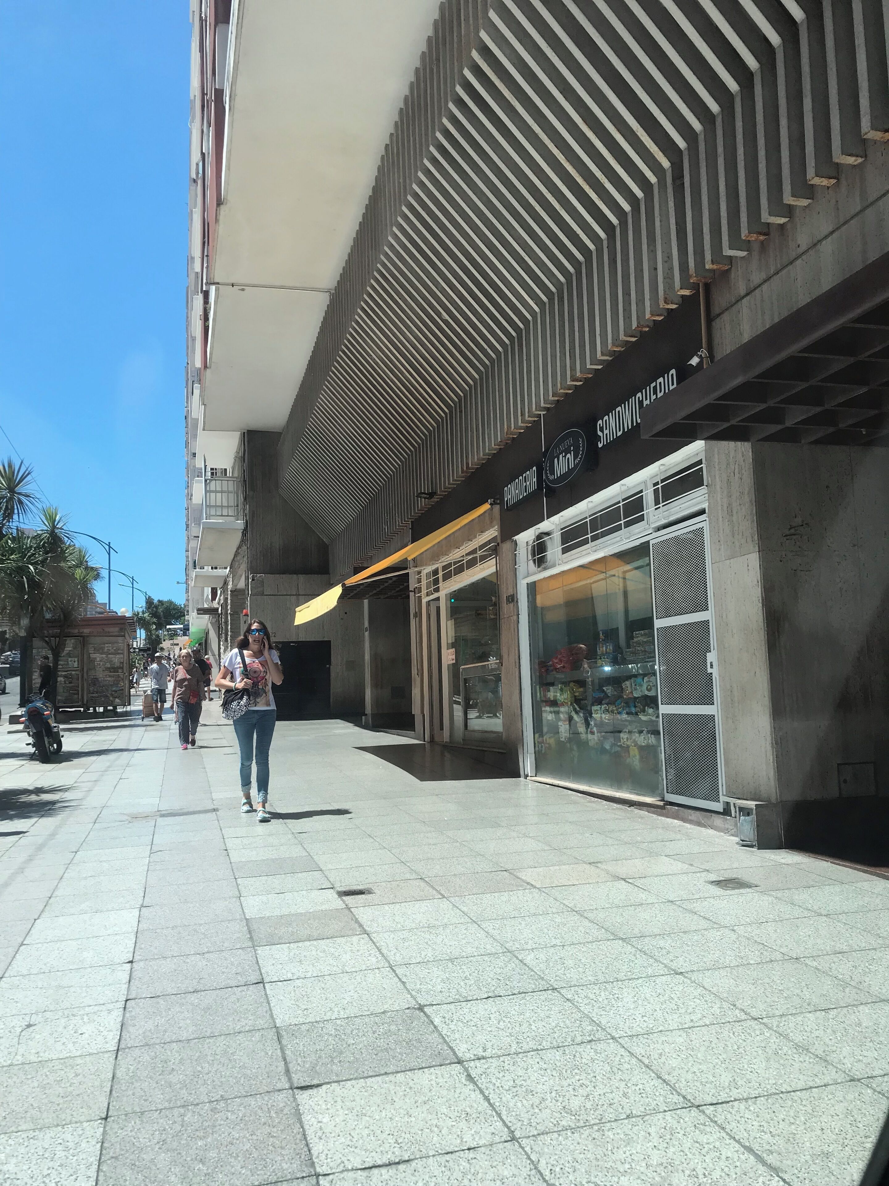 A pedestrian walkway with a modern building featuring a geometric facade and a sandwich shop.