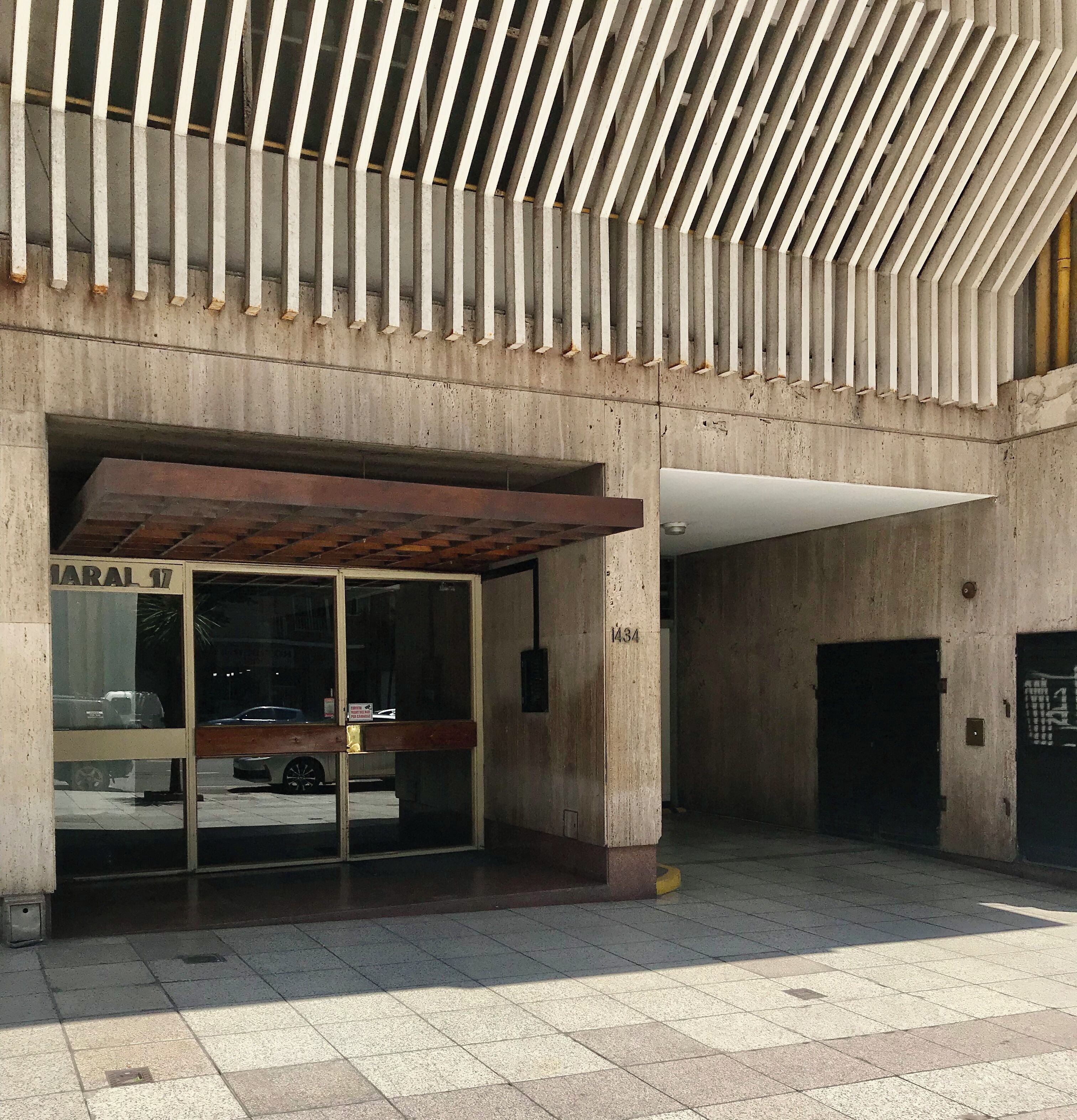 Una entrada de edificio con un gran ventanal, un toldo metálico y fachada de hormigón.