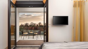 Ein Schlafzimmer mit einem Bett, einem an der Wand montierten Fernseher und einer Glastür, die auf eine Außenpatio mit Stühlen und einem Tisch führt.
