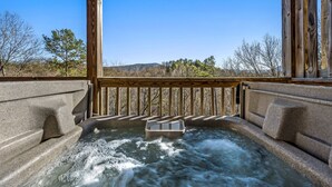 A hot tub with a wooden deck and stone walls, surrounded by a natural landscape with trees and hills. - 20-Mile View, Hot Tub, Game Room, Waterfall (Sevierville)