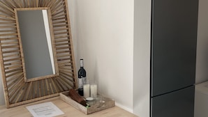 A small kitchenette with a wooden table, two stools, a mirror, a refrigerator, and a bottle of wine.