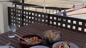 A table with food and drinks under an umbrella, overlooking a beach with palm trees and people in the distance.