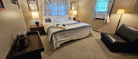 Une chambre d’hôtel comprenant un lit, deux lampes de chevet, un ventilateur de plafond, une fenêtre avec des rideaux, un fauteuil en cuir noir et une petite table avec une lampe.