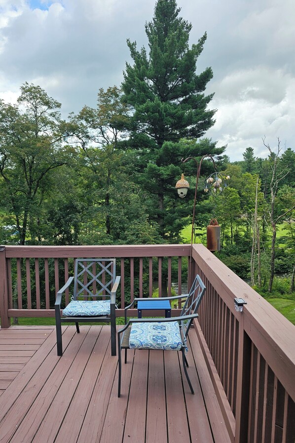 A wooden deck with two chairs and a table, surrounded by a railing, overlooking a forested area.