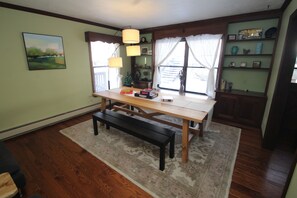 A dining area with a wooden table, benches, and a rug. There are windows with curtains, a shelf with books, and a framed picture on the wall.