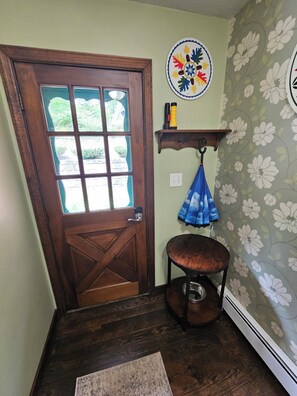 A wooden door with a glass panel, a small wooden stool with a blue cloth, a wall-mounted shelf with a decorative plate, and a patterned wallpaper.