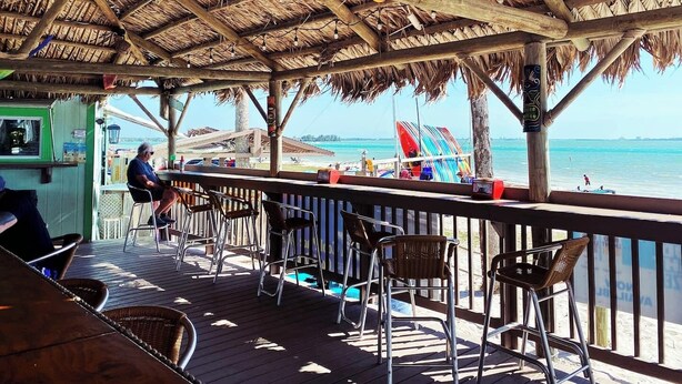 Un espace extérieur abrité, avec des chaises et des tables en bois, donnant sur une plage où l’on peut voir des planches de surf et offrant une vue dégagée sur l’océan.