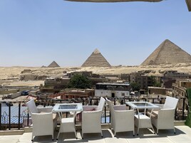 A terrace with wicker furniture and a glass table, overlooking the pyramids and a desert landscape.