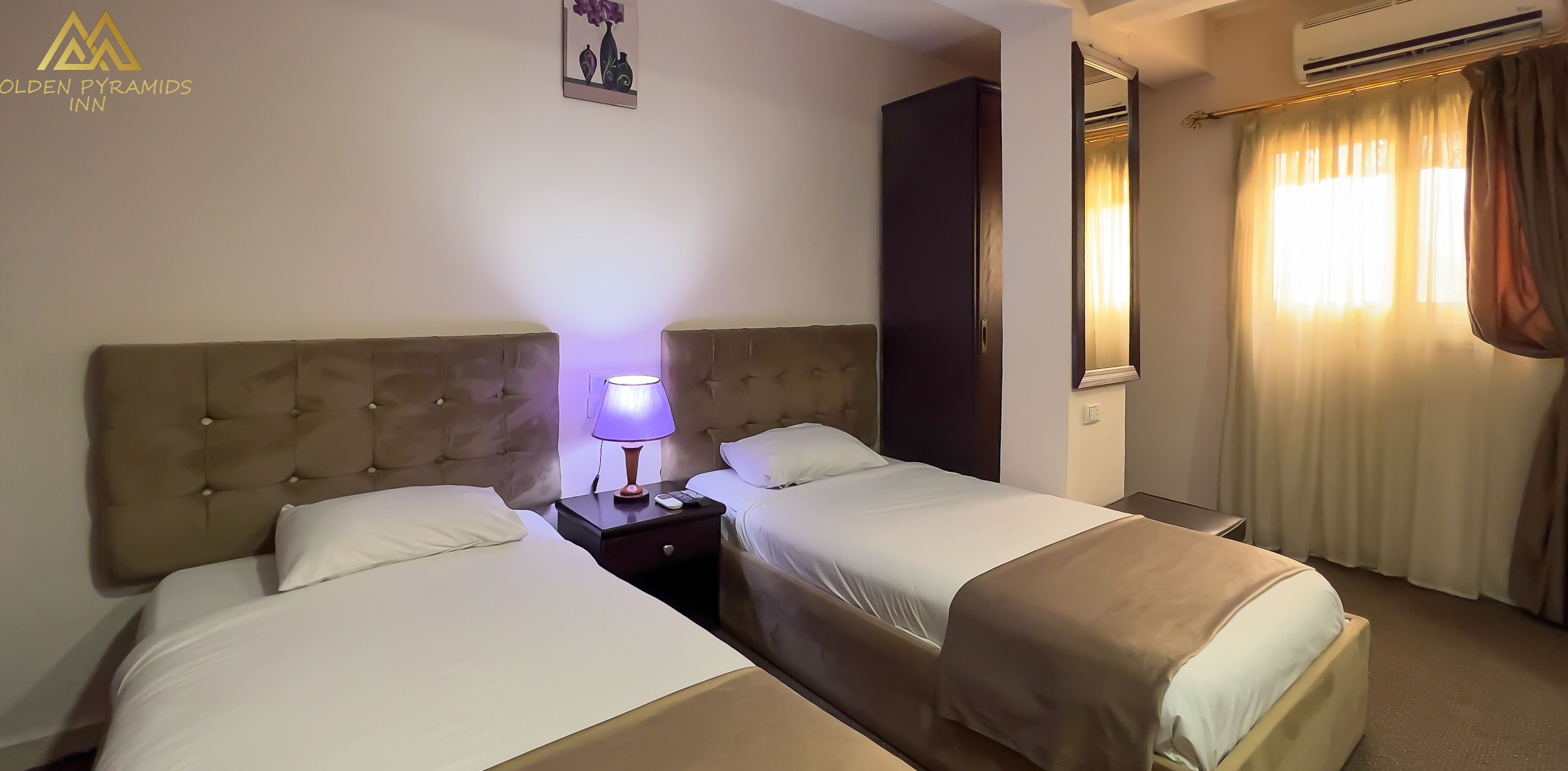 A hotel room with two beds, a nightstand with a lamp, a mirror, and a window with curtains.