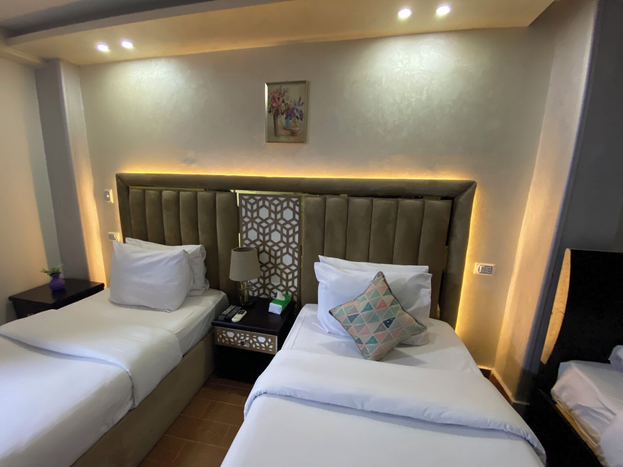 A hotel room with two beds, a headboard with a geometric pattern, a nightstand with a lamp, and a wall-mounted painting.