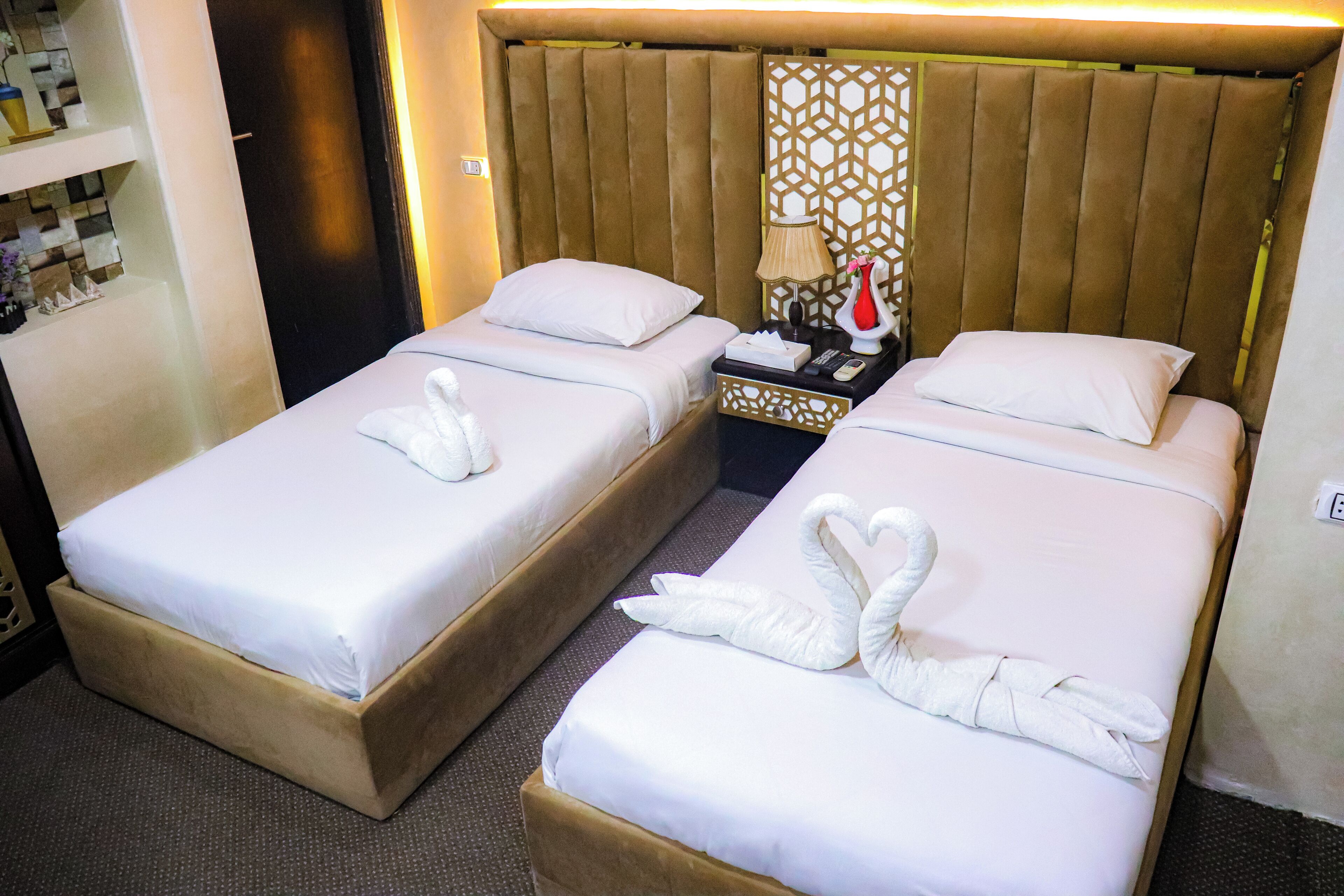A hotel room with two beds, each with a white bedspread and a decorative swan towel. There is a nightstand with a lamp and a vase between the beds.