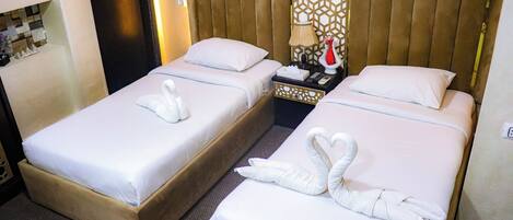 A hotel room with two beds, each with a white bedspread and a decorative swan towel. There is a nightstand with a lamp and a vase between the beds.