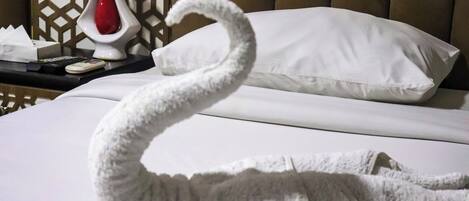 Una toalla con forma de cisne sobre una cama con sábanas blancas, un jarrón rojo y un control remoto en una mesita de noche.