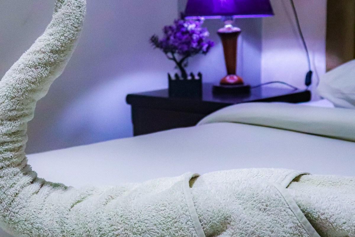 A neatly made bed with a towel shaped like a swan, a purple lamp, and a small vase with flowers on a bedside table.