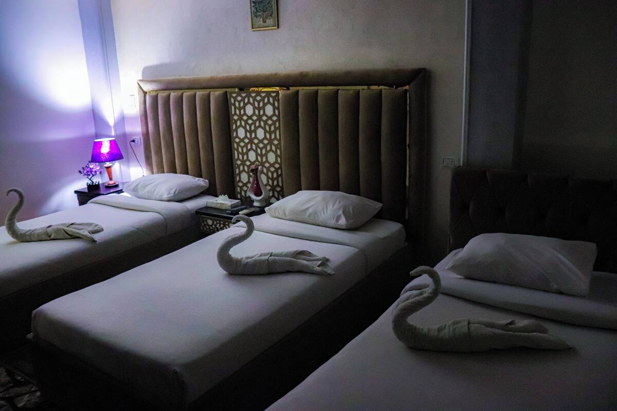 Quarto de hotel com duas camas de solteiro, cada uma com toalha em formato de cisne. Há uma mesa de cabeceira com abajur e um objeto decorativo.