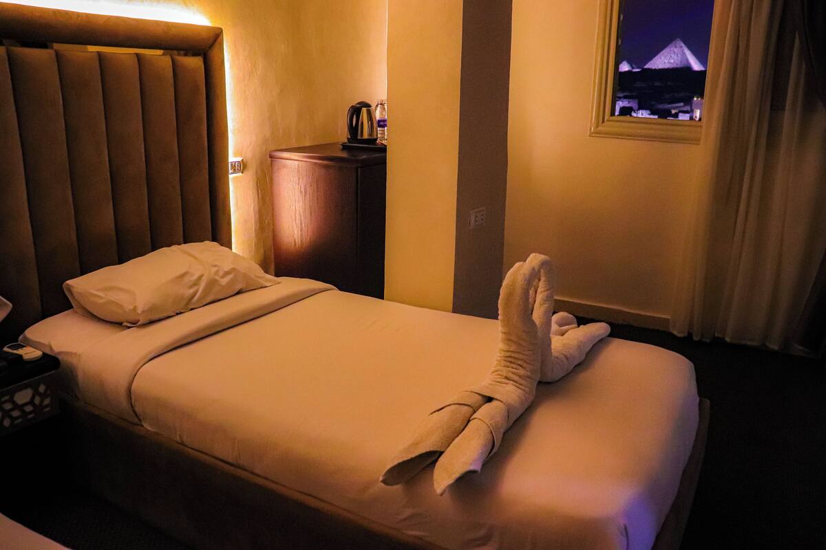 Quarto de hotel com cama, mesa de cabeceira, luminária, janela com vista para uma pirâmide e toalha sobre a cama.