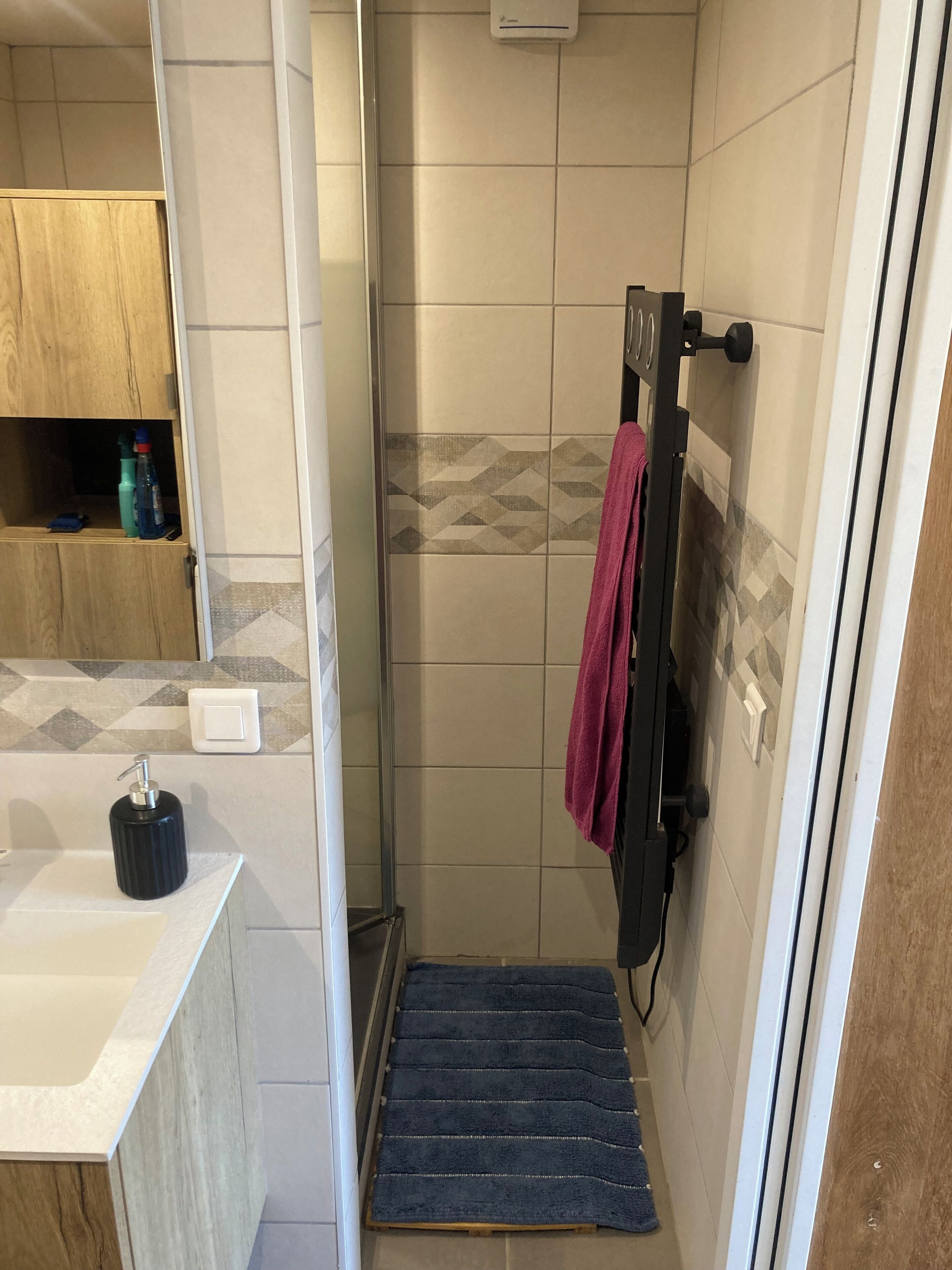 A bathroom with a shower, a wooden cabinet, a black soap dispenser, and a towel rack with a red towel.