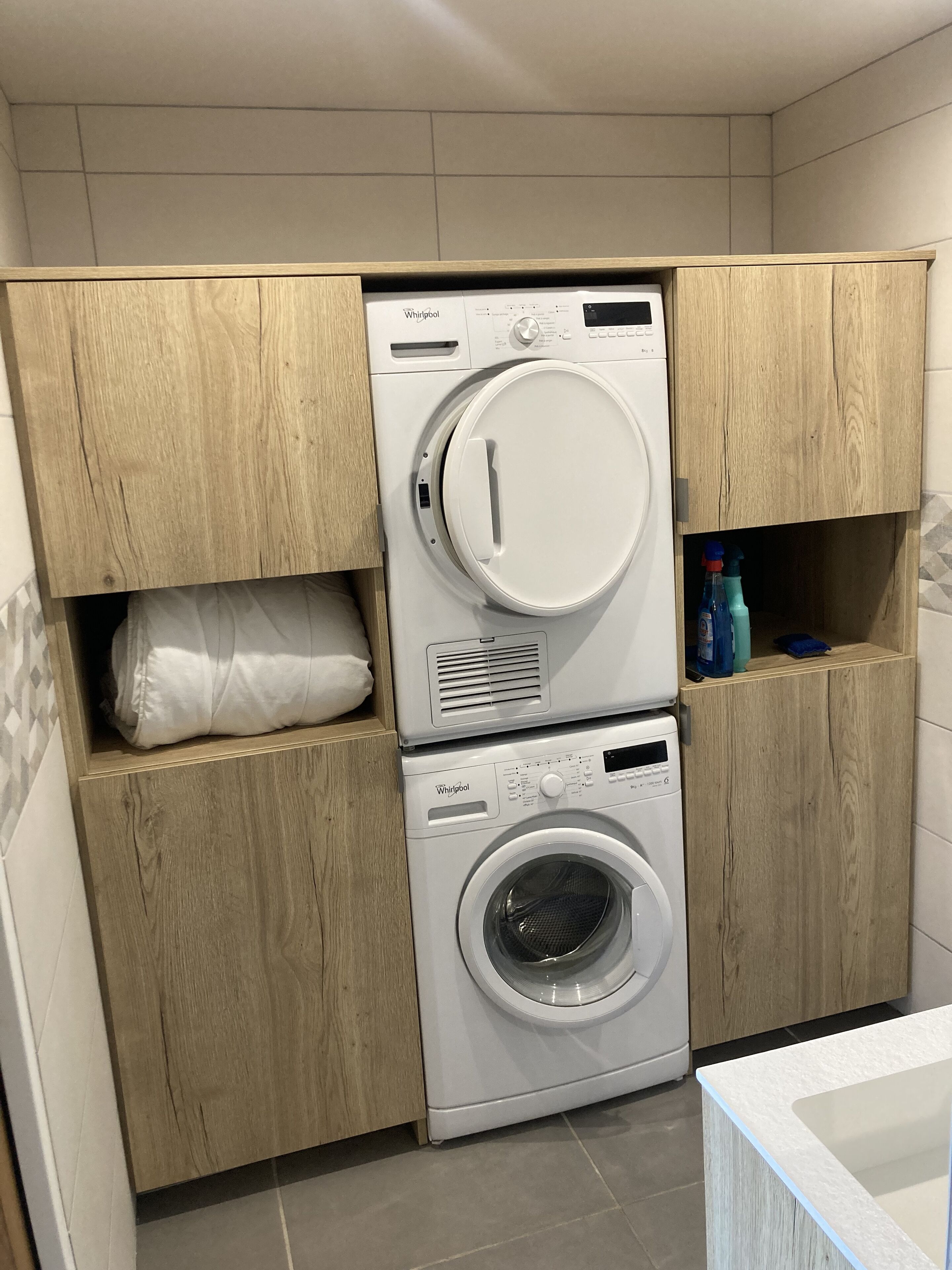 Ein Waschraum mit zwei übereinander gestapelten Whirlpool Waschmaschinen und Trocknern, Holzschränken und einem Regal mit Reinigungsmitteln.