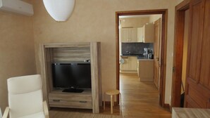 Una sala de estar con televisor, una silla, un taburete pequeño y un mueble de madera. Se ve una cocina al fondo.