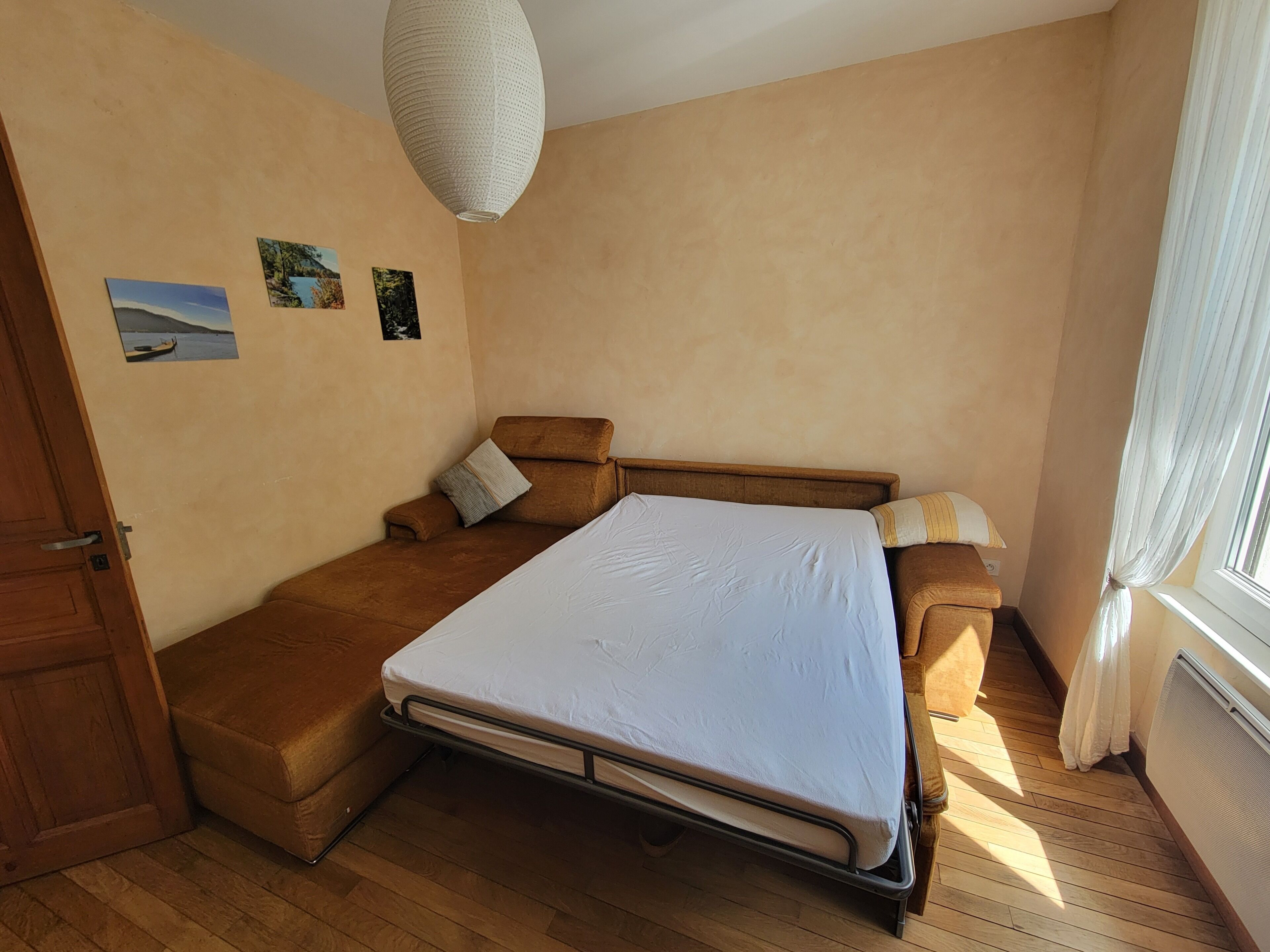 Un dormitorio con una cama, un sofá, piso de madera, una lámpara de techo y una ventana con cortinas.