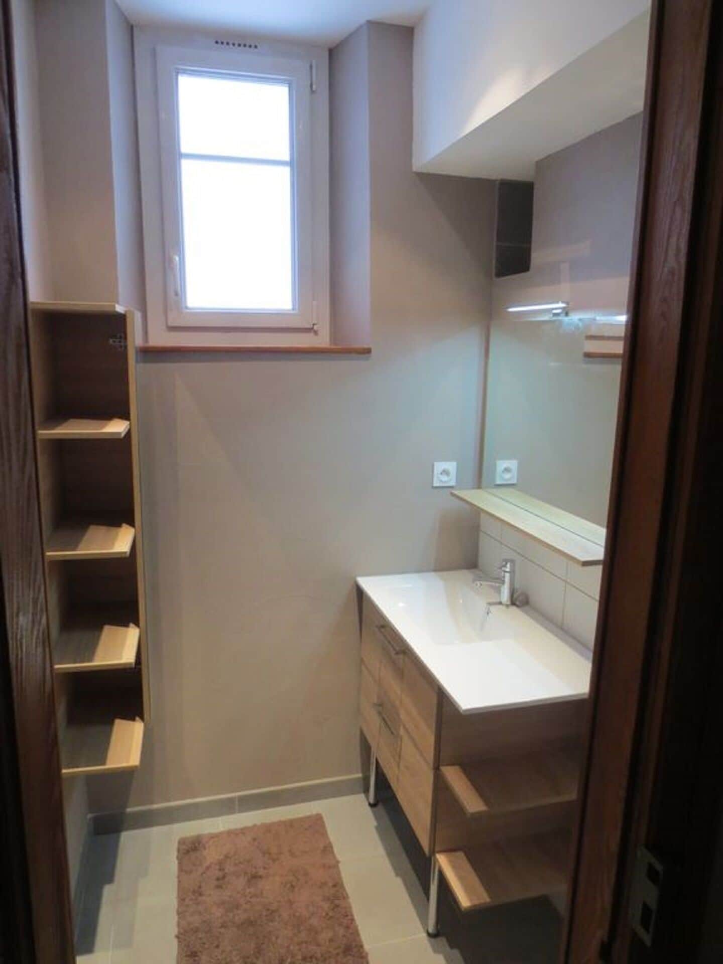 Een kleine, smalle badkamer met een raam, een witte wastafel, een spiegel en een houten wastafelmeubel.