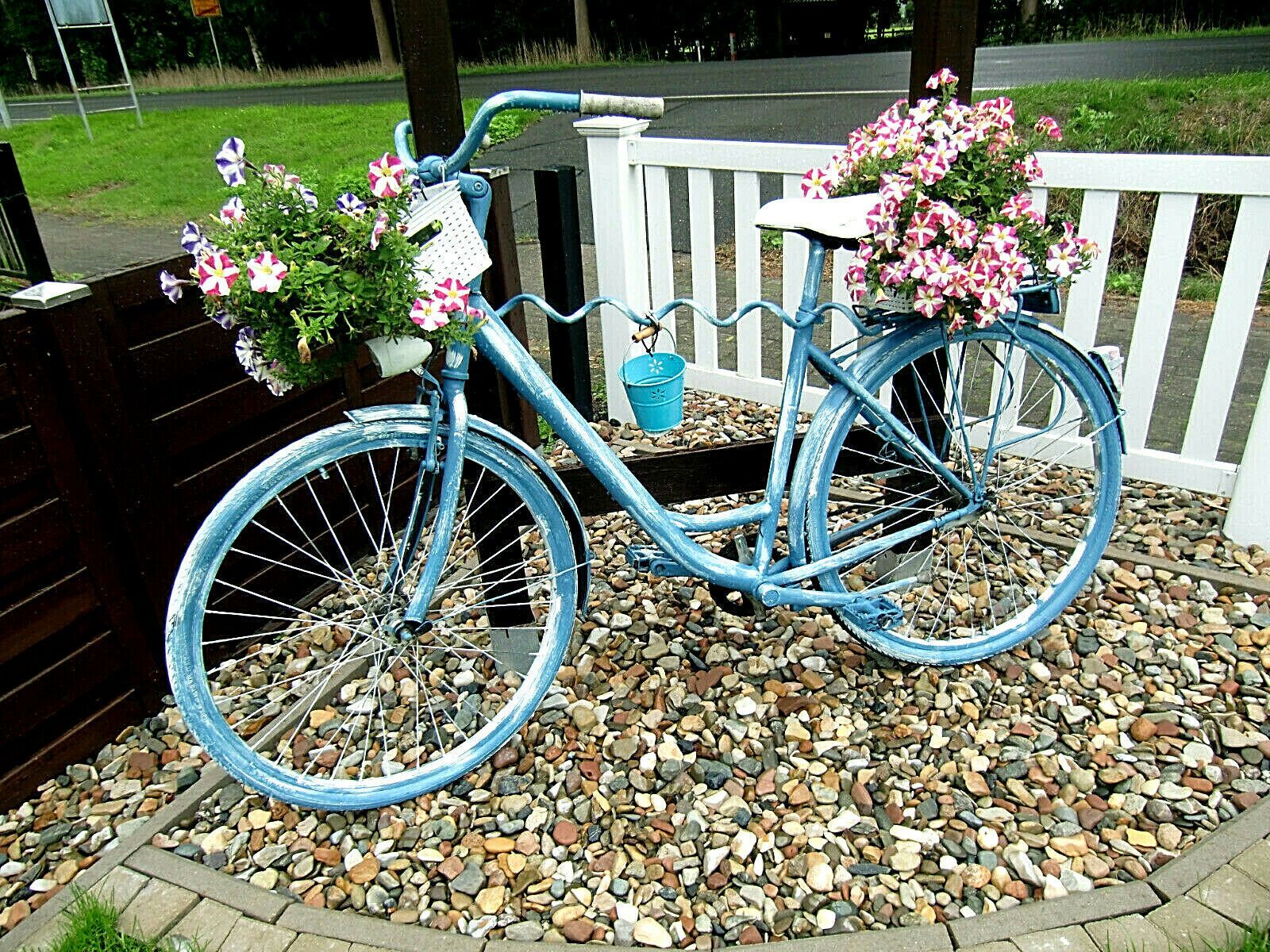 Ein blaues Fahrrad mit Korb und Blumen, abgestellt auf einem Schotterweg mit weißem Zaun und Grünpflanzen im Hintergrund.