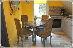 A dining area with a glass-top table and wicker chairs, a kitchen with a coffee maker, and a clock on the wall.