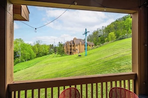A view from a balcony with a wooden railing, overlooking a grassy hill with a building and a ski lift in the background. - SnowPine Condo, Sleeps 8, A/C, Fireplace (Ellicottville)