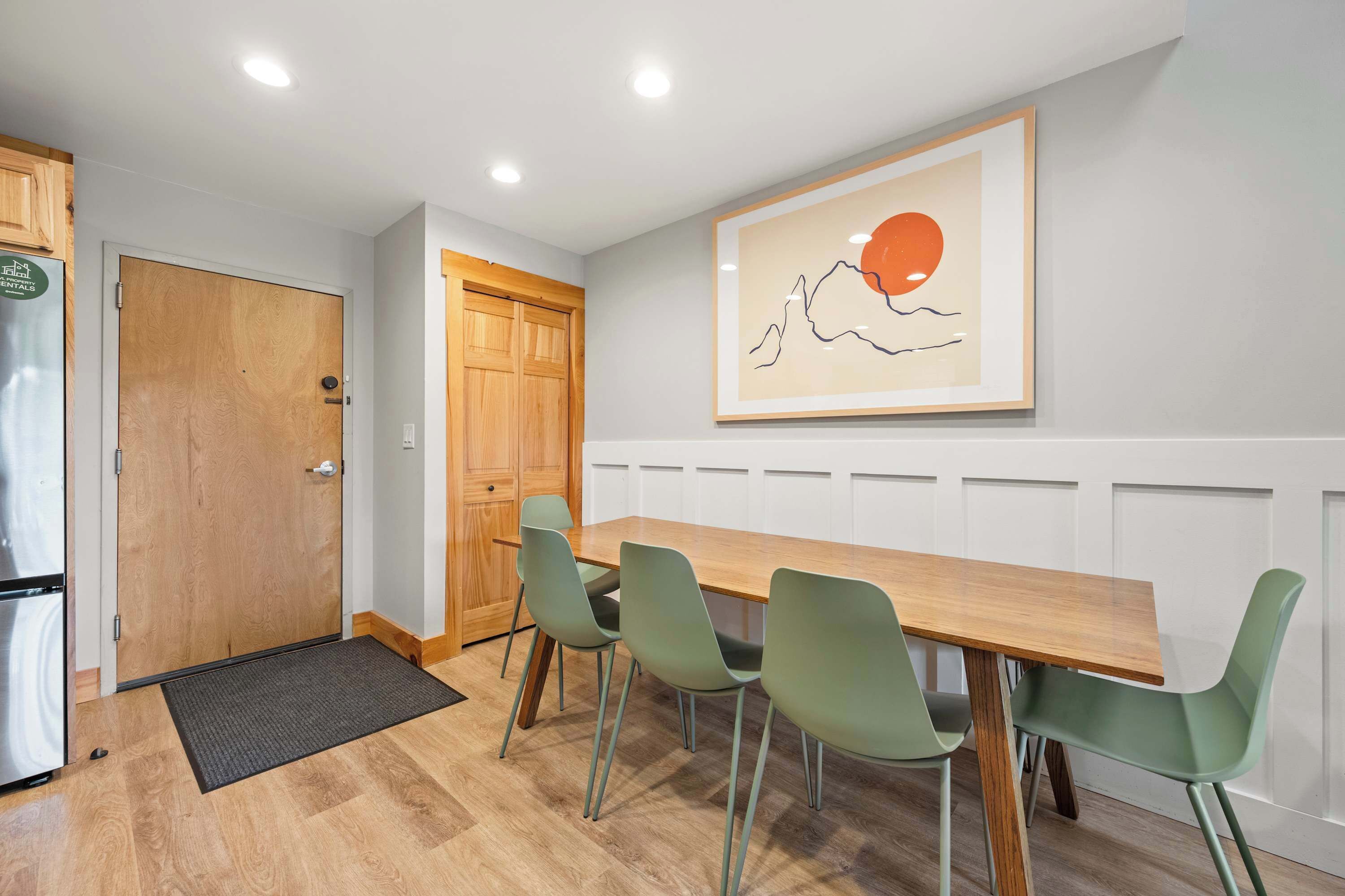 A dining area with a wooden table and four green chairs, a wooden door, and a wall-mounted artwork.