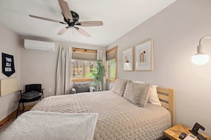 Ein Schlafzimmer mit einem großen Bett, einem Deckenventilator, einem Schreibtisch mit Stuhl, einem Fenster mit Vorhängen und einer Wand-Klimaanlage.