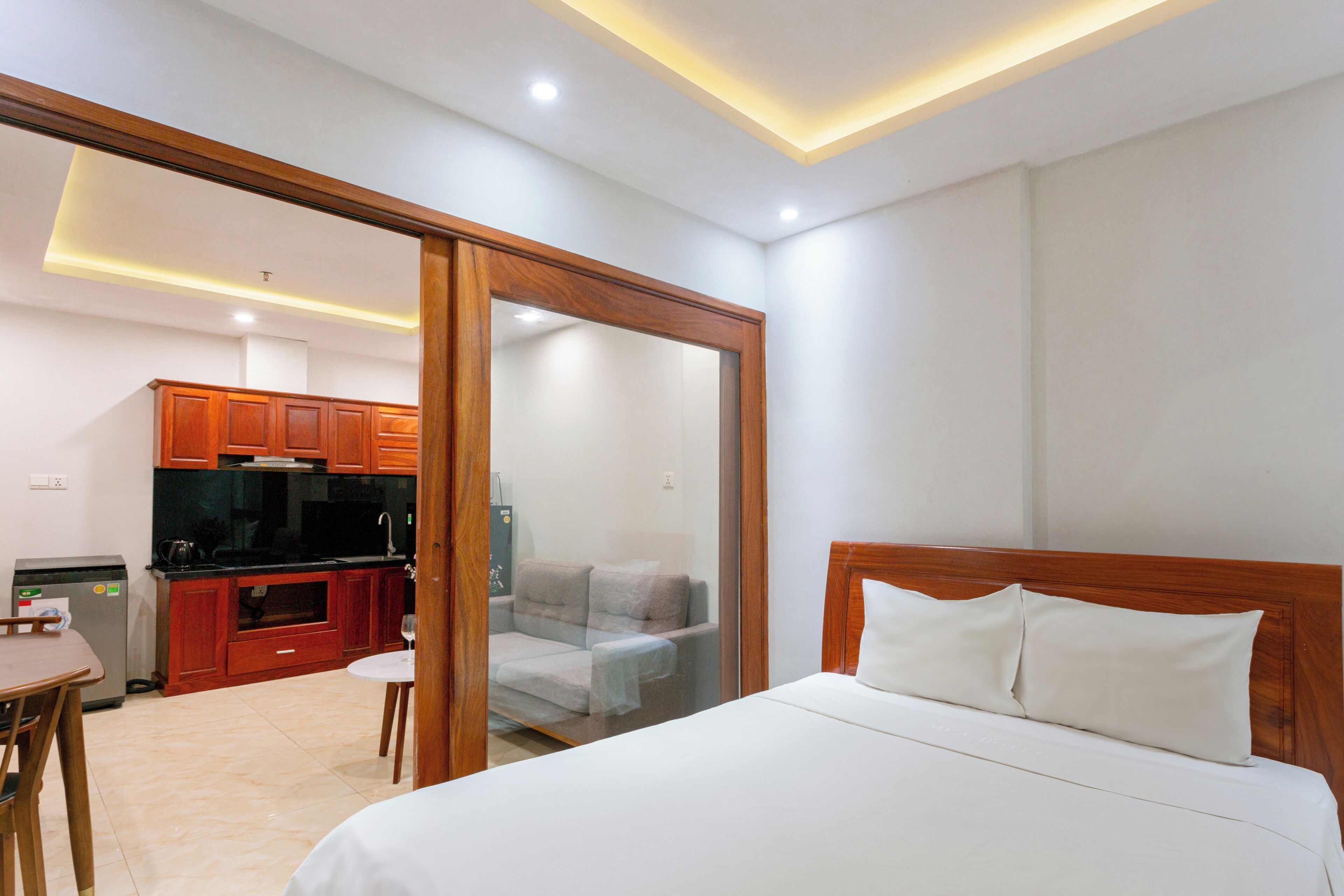 Ein Schlafzimmer mit einem Bett, einem Holz-Kopfteil und Blick in einen Küchenbereich mit Schränken und einem Kühlschrank.