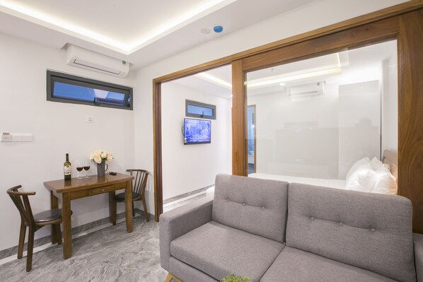 A modern living room with a grey sofa, wooden dining table, and chairs. There is a TV on the wall and an air conditioning unit above it.