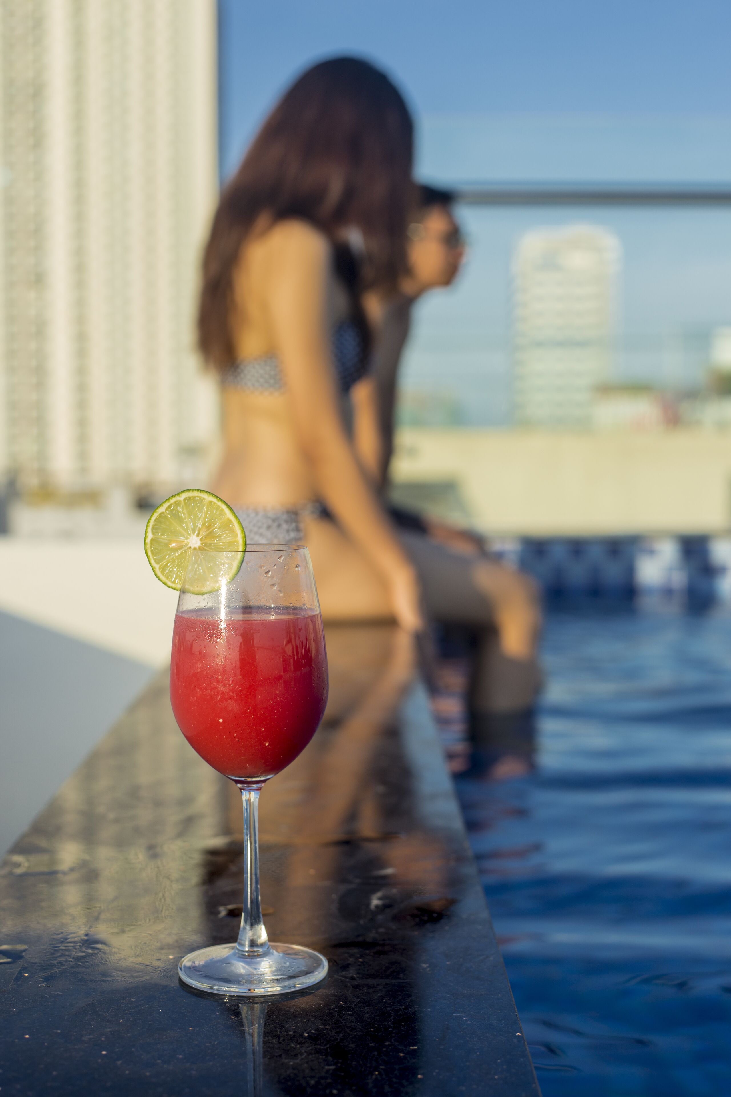 A glass of red drink with a lime slice on a reflective surface near a poolside with a person in the background.