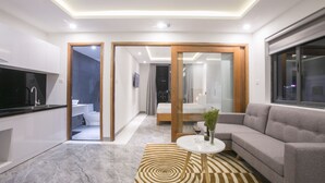 Una sala moderna con un sofá, una mesa de centro y un televisor. Se observa una zona de cocina con armarios y una encimera. La habitación tiene un ventanal grande y una puerta que da a otra estancia.