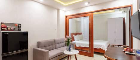 A hotel room with a sofa, a small table with a vase, a bed, a TV, and a refrigerator.
