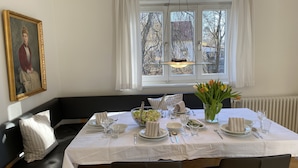 Une salle à manger avec une table dressée pour quatre personnes, un banc en cuir noir, une fenêtre avec des rideaux et un tableau accroché au mur.