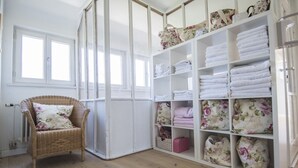 Une pièce avec un fauteuil en osier, une étagère blanche où sont pliés des serviettes, et une fenêtre avec des stores.
