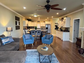 Ein geräumiges Wohnzimmer mit einem grauen Sofa, zwei blauen Sesseln und einem Couchtisch. Eine Deckenventilator und eingelassene Beleuchtung sind zu sehen.