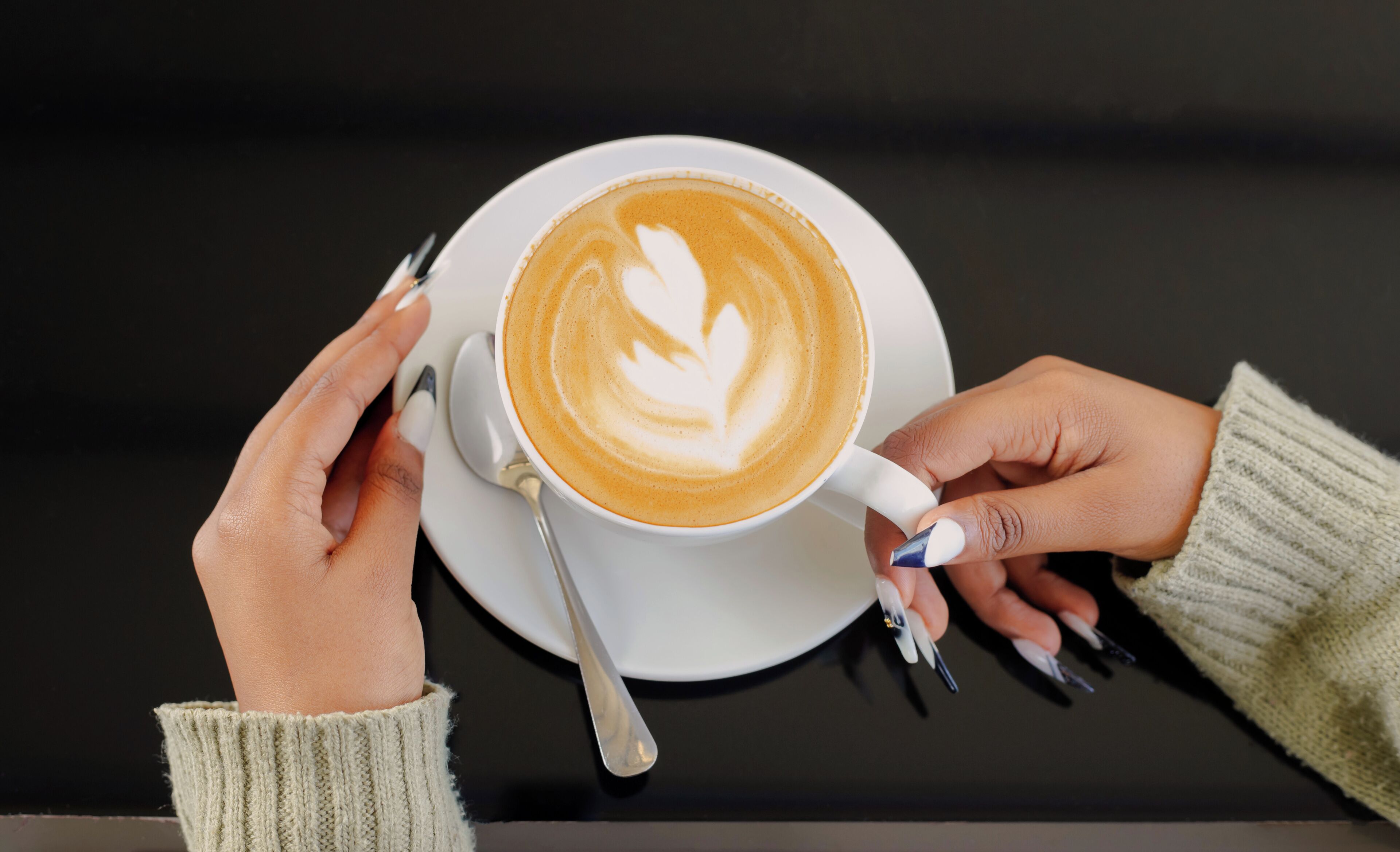 Een kop koffie met latte-art op een schotel, vastgehouden door twee handen.