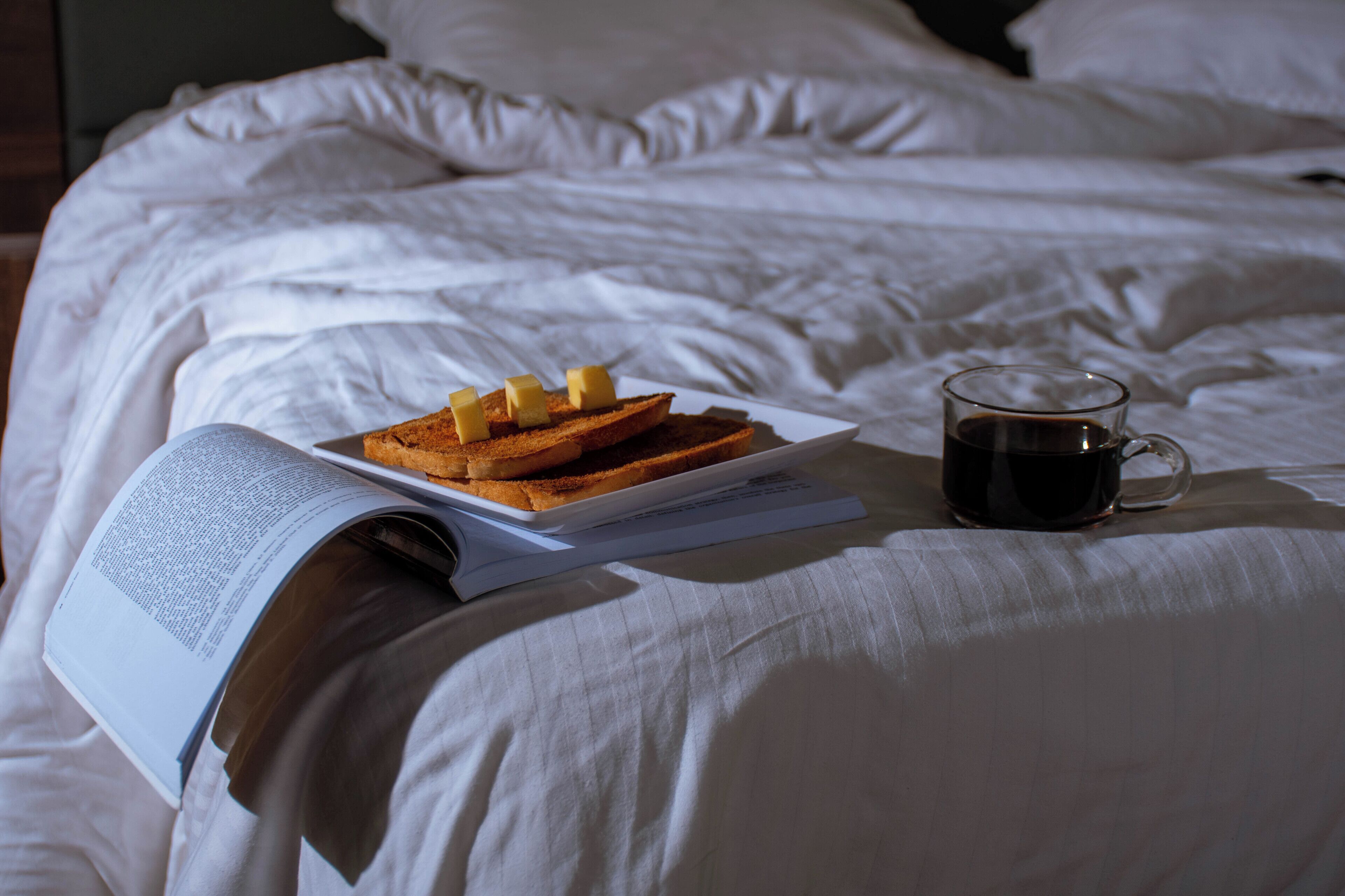 A bed with a book, toast with butter, and a cup of coffee.
