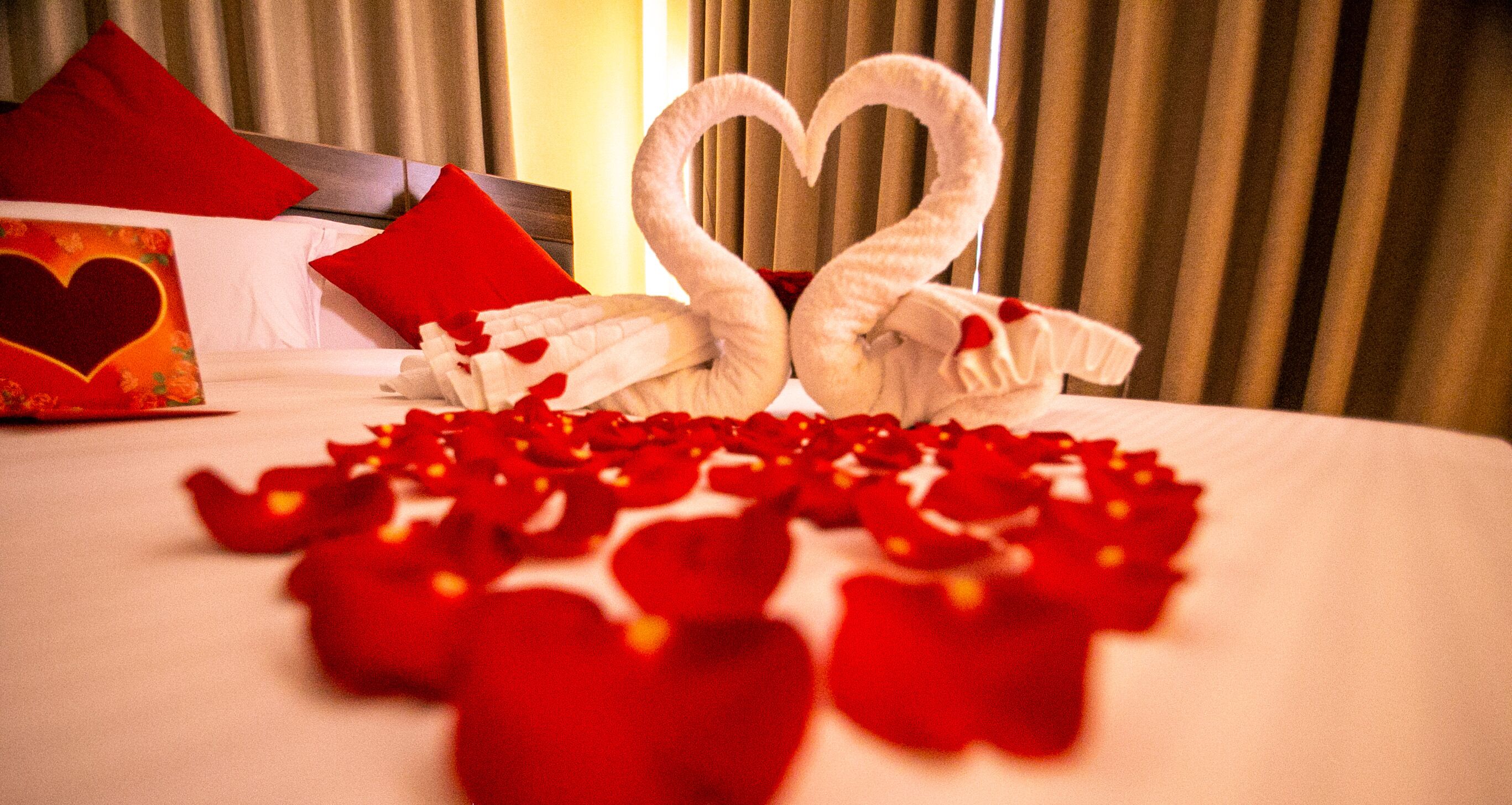 A bed with a heart-shaped towel arrangement and scattered rose petals.