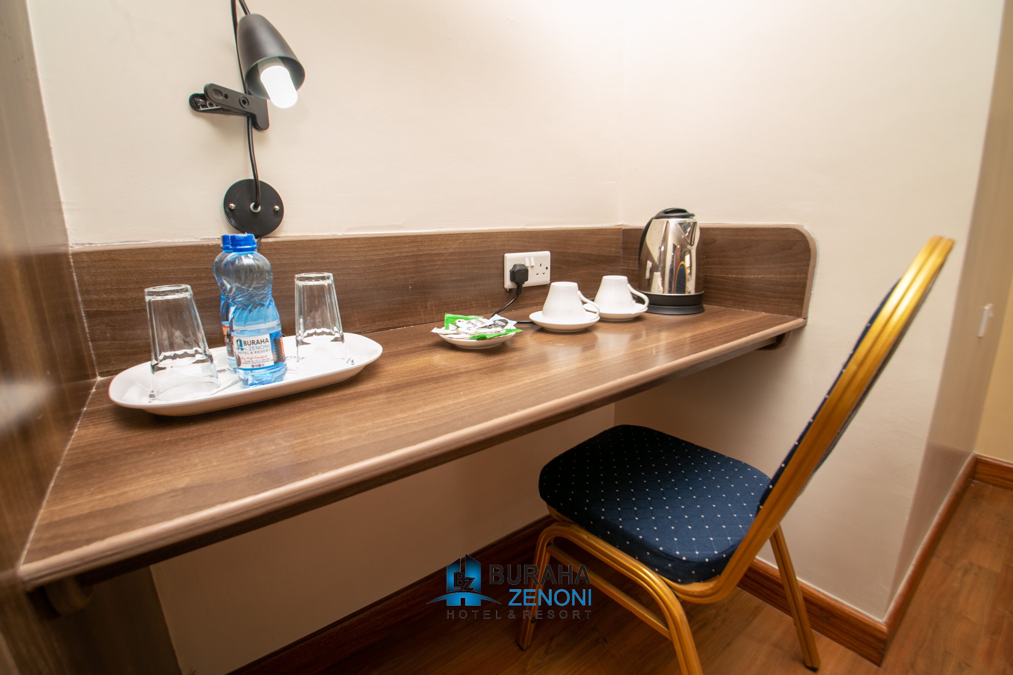 A hotel room with a wooden counter, a chair, a water bottle, glasses, a plate with food, a cup, and a kettle.