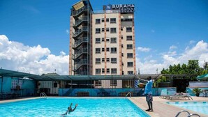 A swimming pool with a person diving, a building labeled "BURAHA ZENONI RESORT" in the background, and a covered area with tables and chairs.