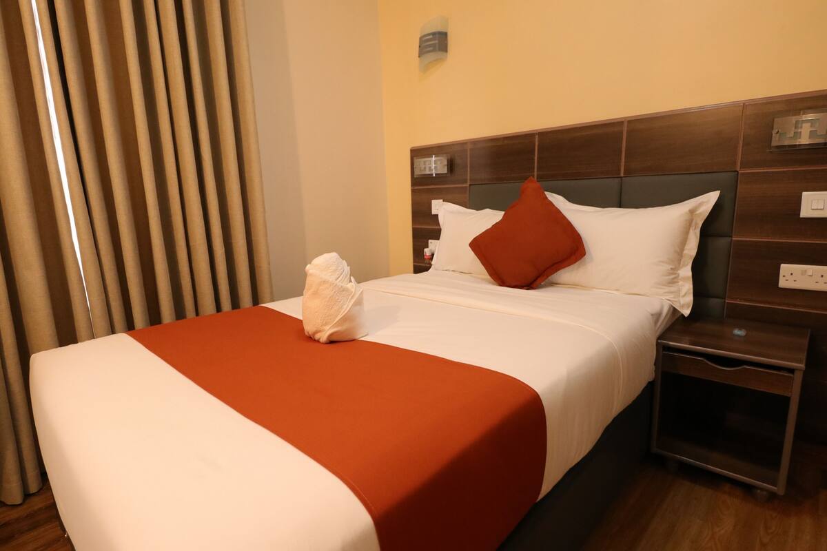 A hotel room with a bed, a nightstand, a wall-mounted light, and a window with curtains.