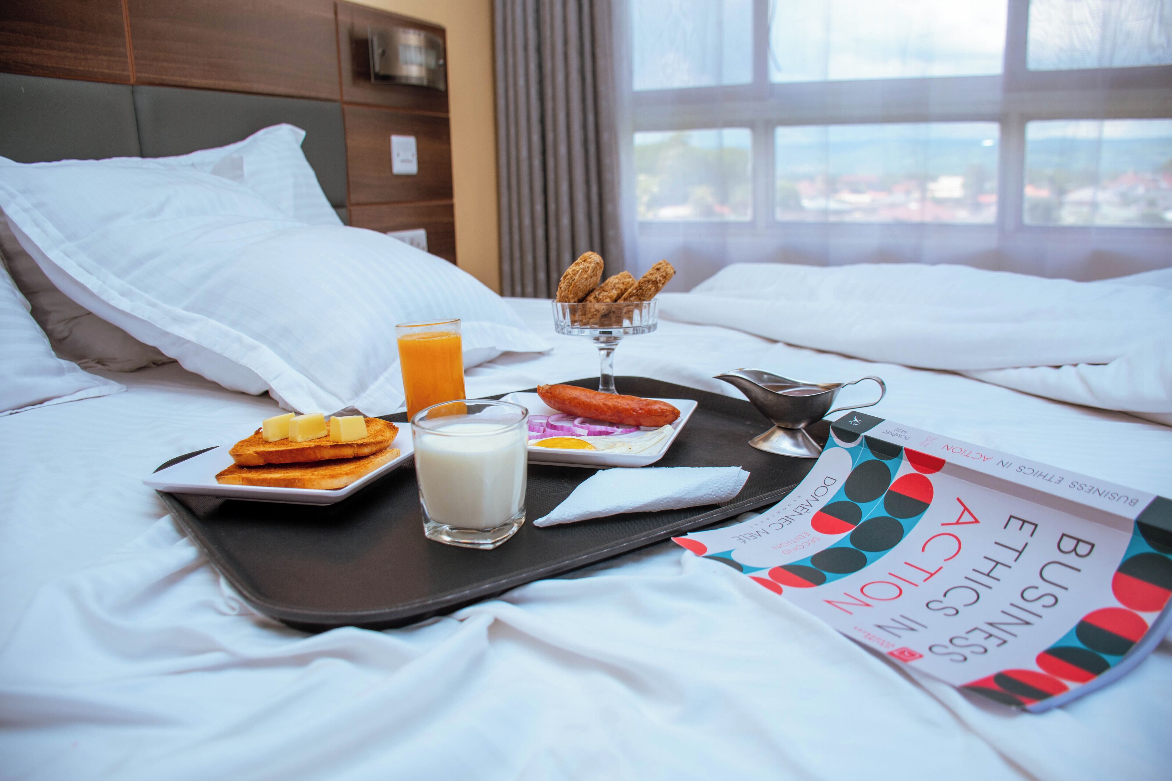 A neatly made bed with white linens, a tray with breakfast items including toast, a glass of orange juice, and a small bowl of cereal.