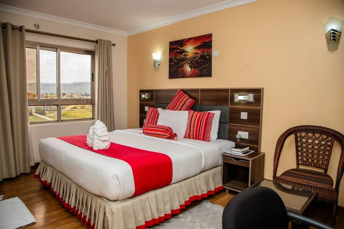 A hotel room with a large bed, a view of the countryside, a painting on the wall, and a wicker chair.