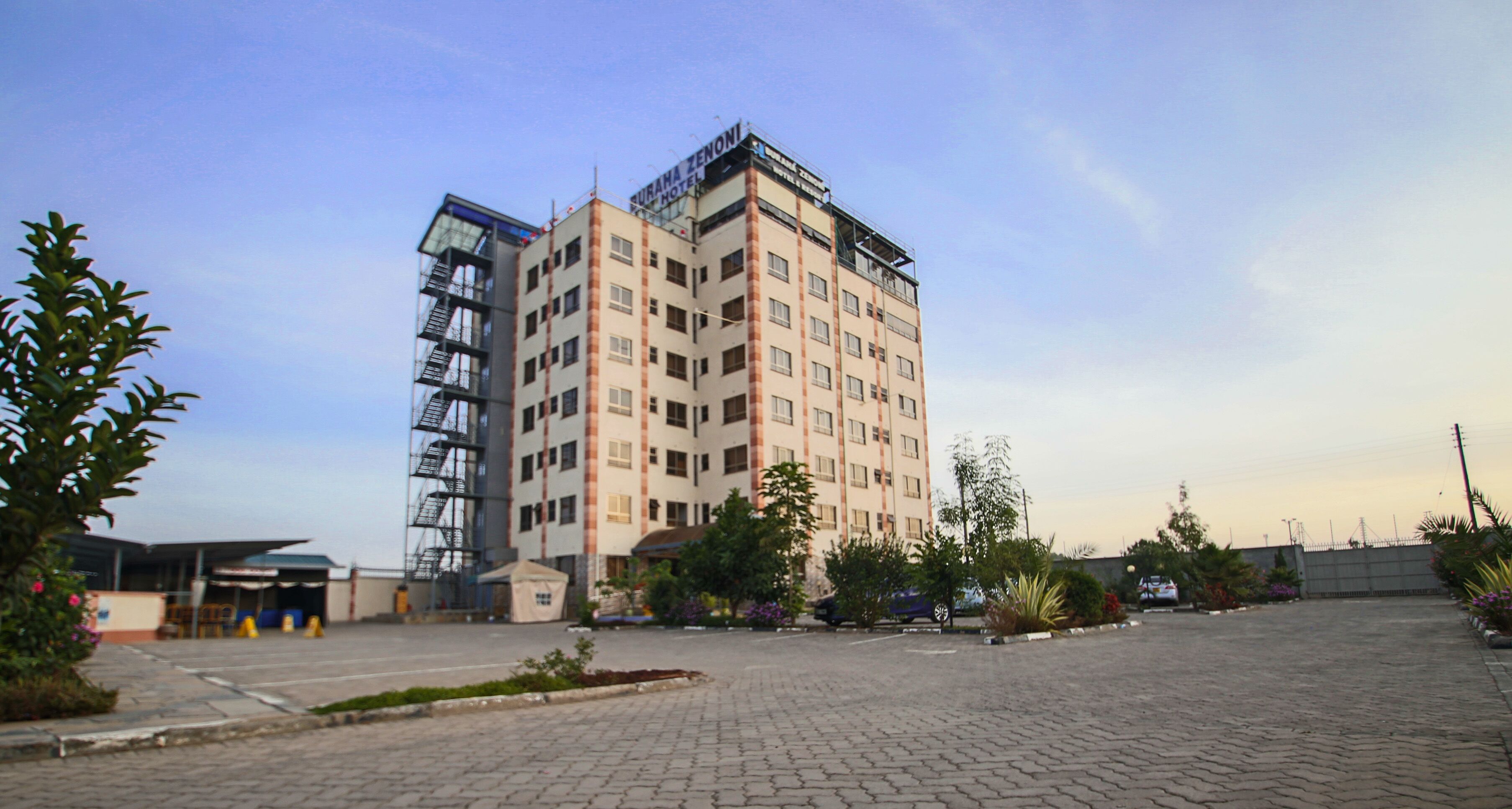 A multi-story hotel with a modern design, featuring a rooftop terrace and a parking area with a few cars.