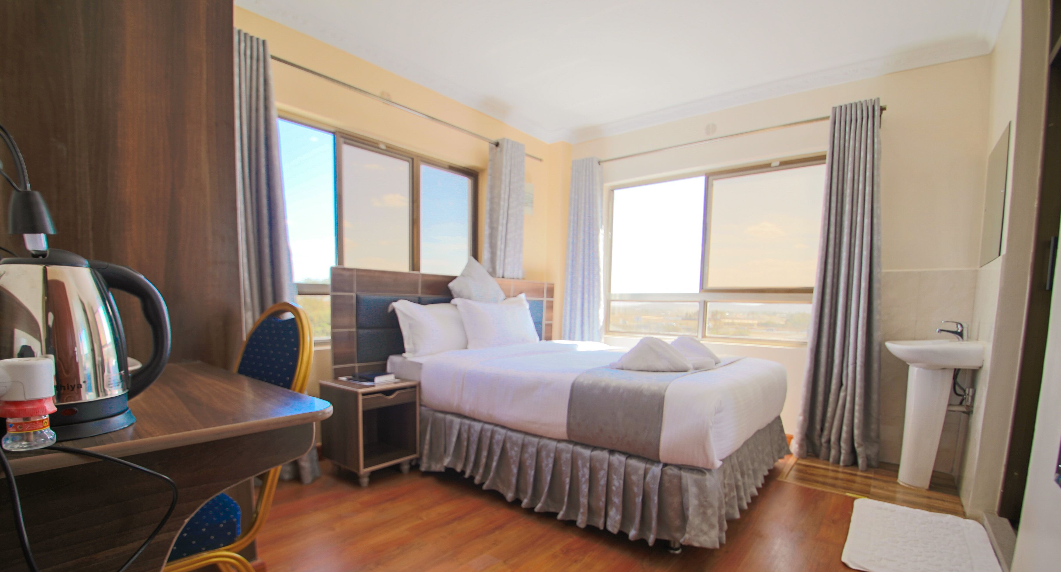 Habitación de hotel con una cama grande, un escritorio con hervidor, una silla, una mesita de noche y vistas a la ciudad desde las ventanas.