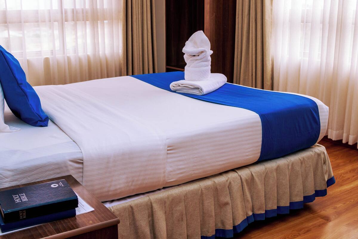 A neatly made bed with a blue and white bedspread, a folded white towel, and a book titled "HOLY BIBLE" on a wooden nightstand.